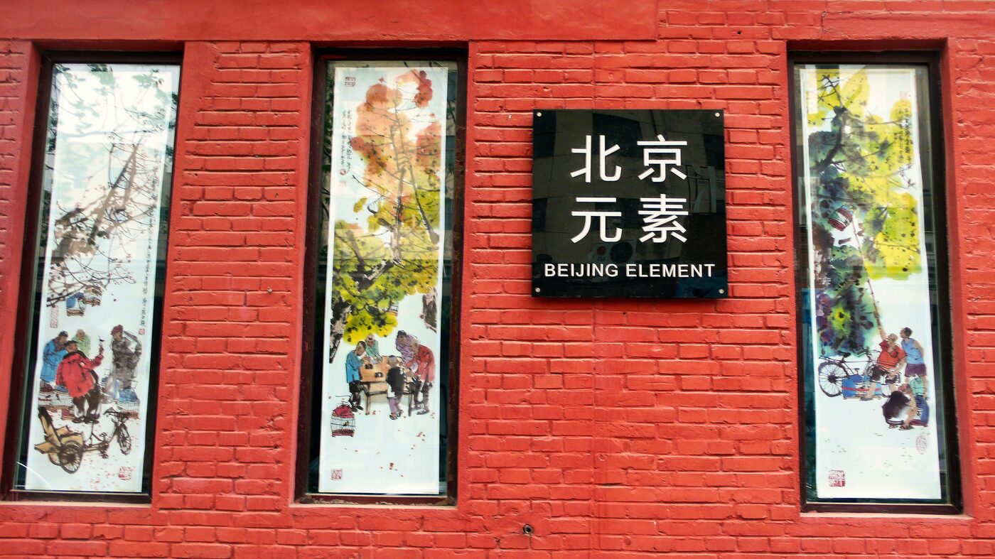 A red-brick factory wall hung with framed artworks beside a gallery sign in the 798 Art District, Beijing.