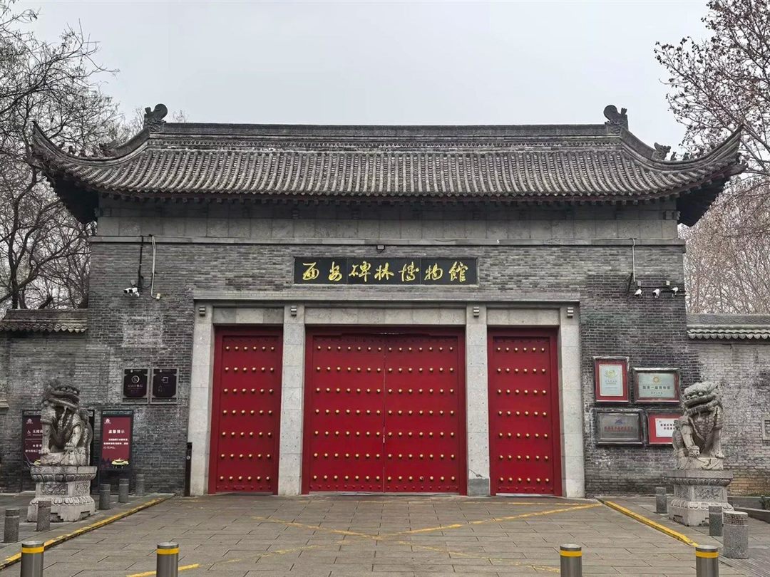 A grey-brick gatehouse under a sweeping tiled roof, with a gold-on-black plaque reading 西安碑林博物館, three studded red doors and a stone lion each side.
