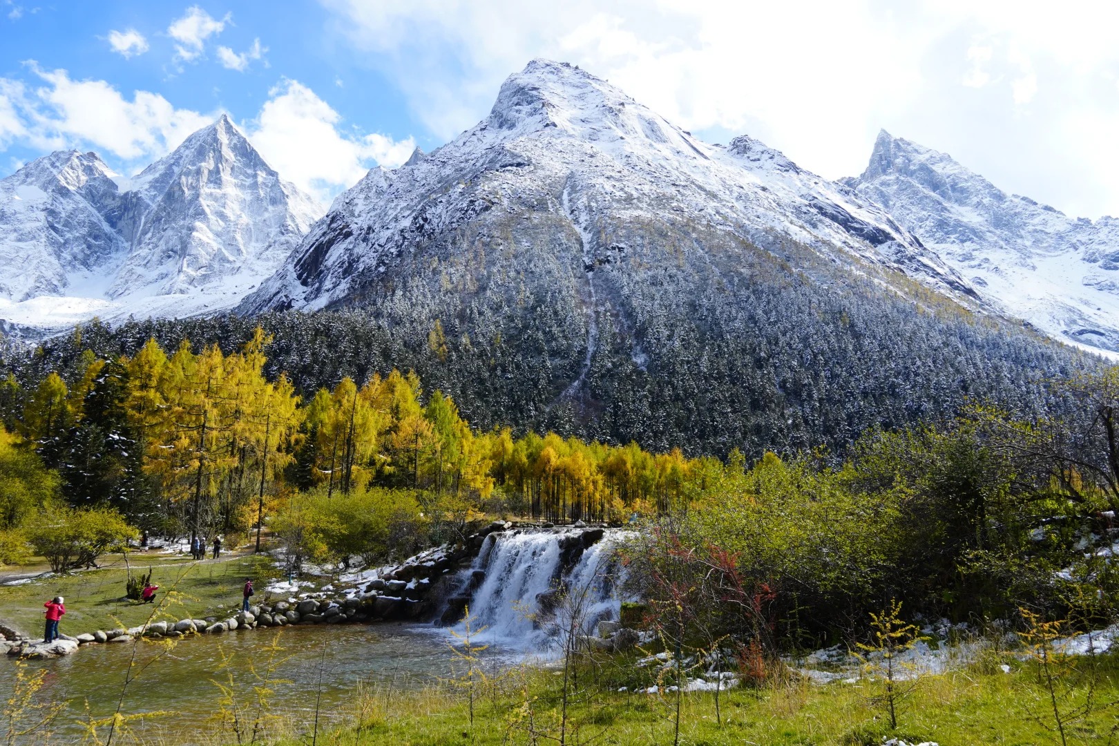 Shallow clear water running over a stone-lined step, golden larch along the banks and a broad snow-covered massif rising behind, with a few visitors standing on the stones.