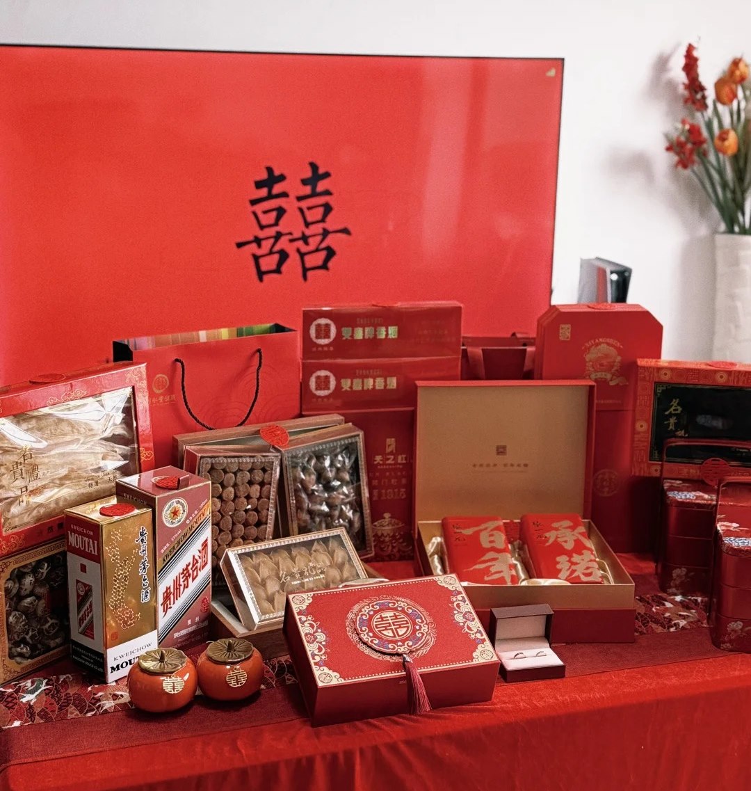 A table of red betrothal gifts laid out: spirits, cigarettes, cake boxes, dried-seafood hampers, tea caddies and an open ring box
