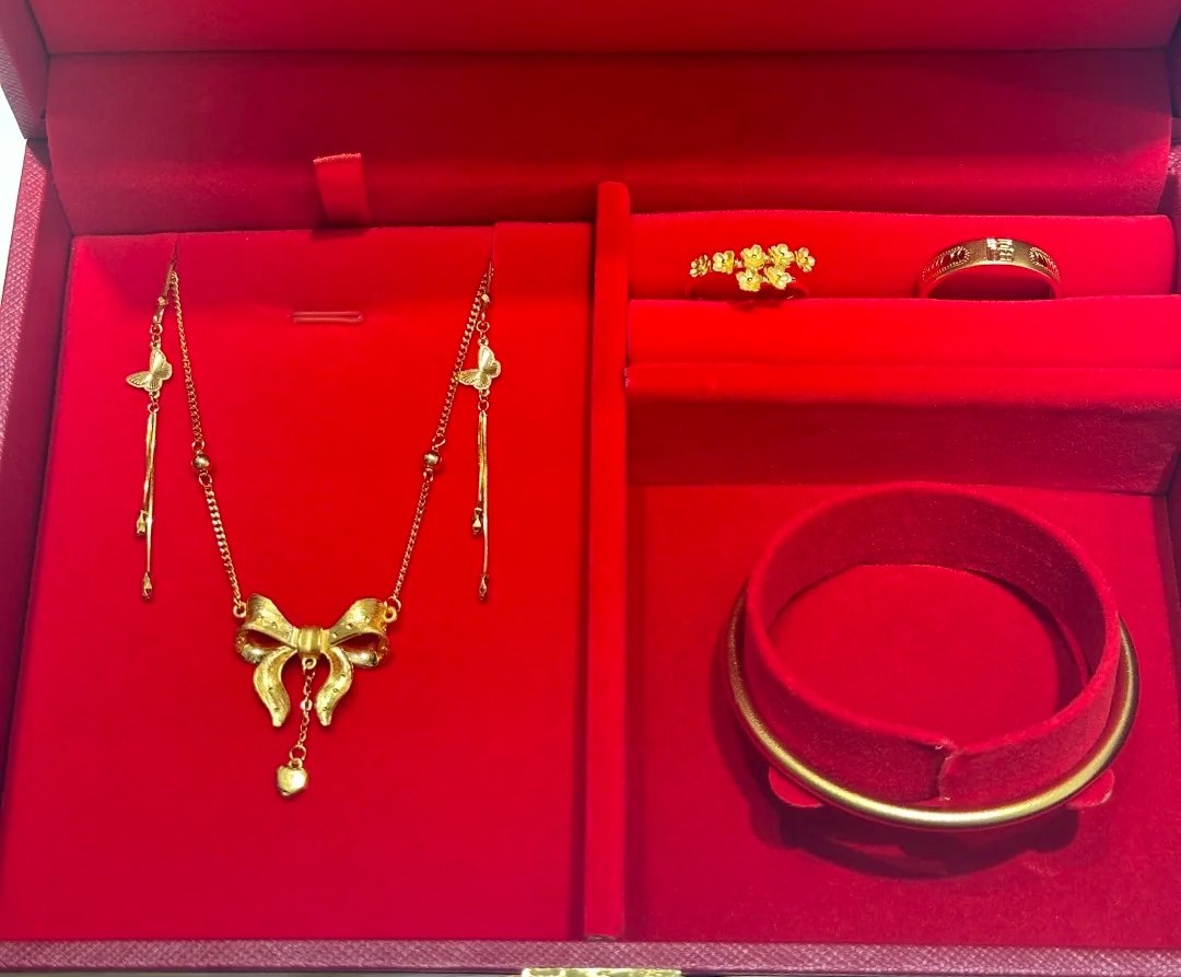 A red velvet presentation box holding a gold necklace with a bow pendant, two gold rings and a gold bangle