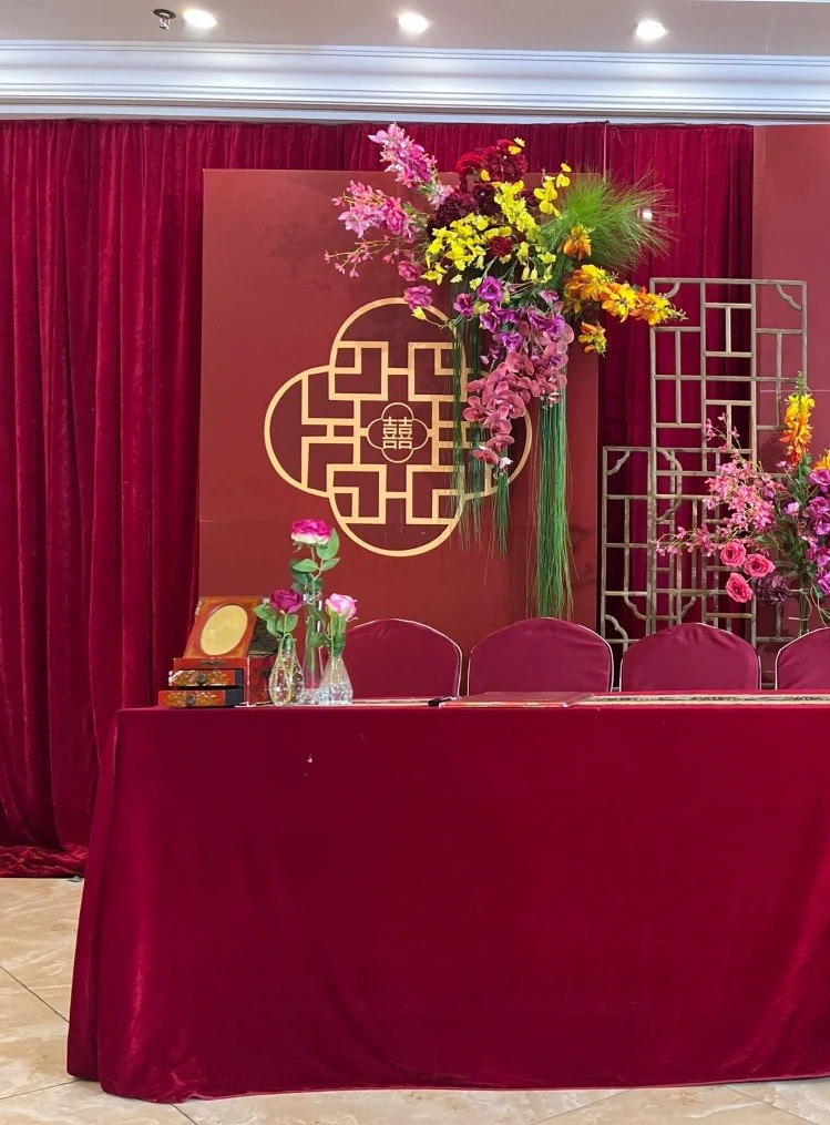 A Chinese wedding sign-in desk in deep red velvet, with a guest register, an ink-seal box and flowers, in front of a double-happiness board