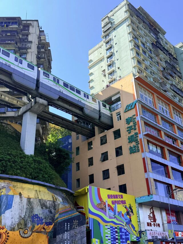 Wider street-corner view of the Liziba monorail station building from the opposite side of the road, showing the canyon-edge geography of the Jialing River bank.