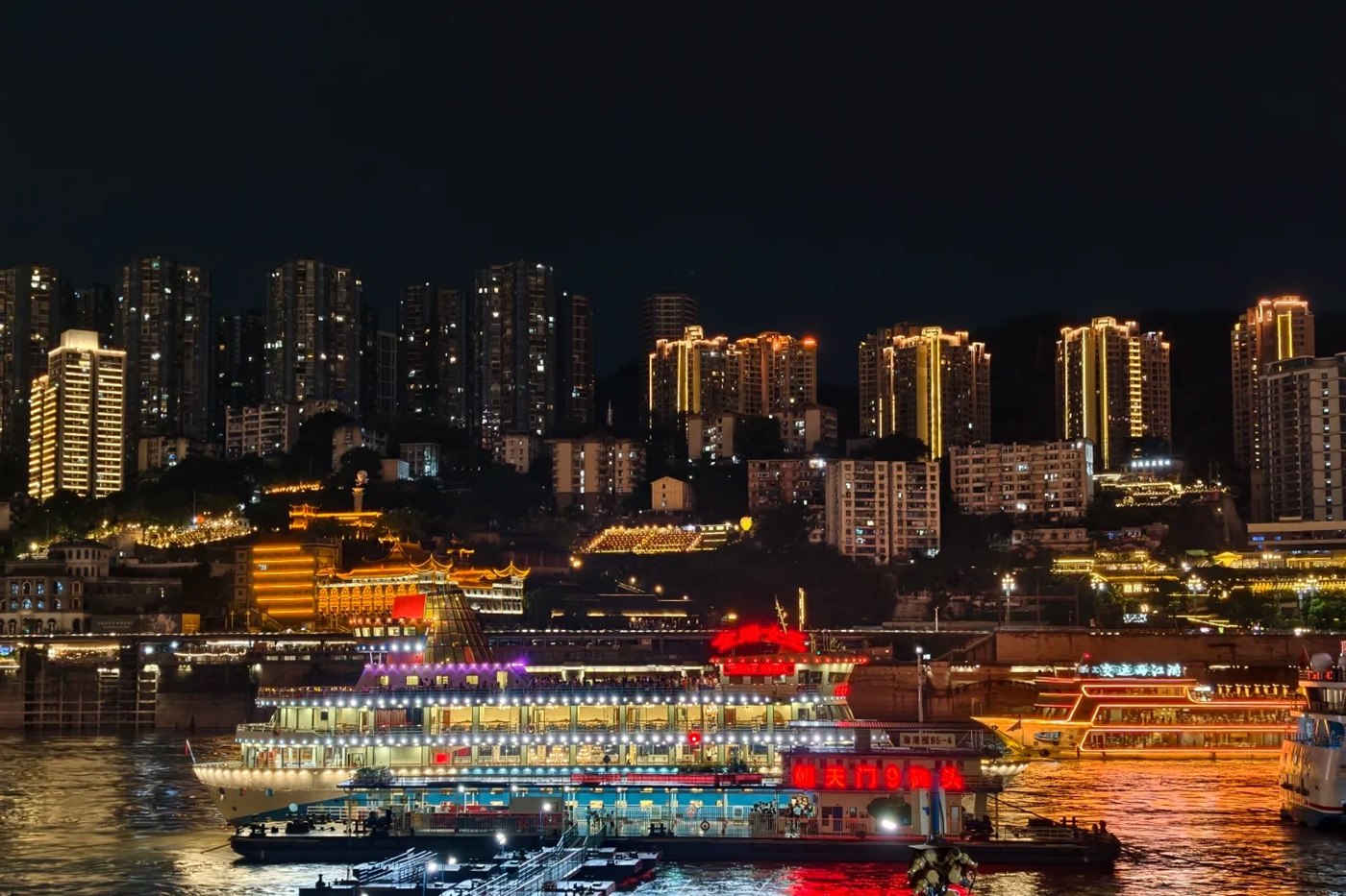 A lit sightseeing boat berthed at a floating pontoon in Chongqing at night, with large red characters reading 朝天门9码头 (Chaotianmen Pier 9) on the pontoon wall, apartment towers stacked up the hillside behind.