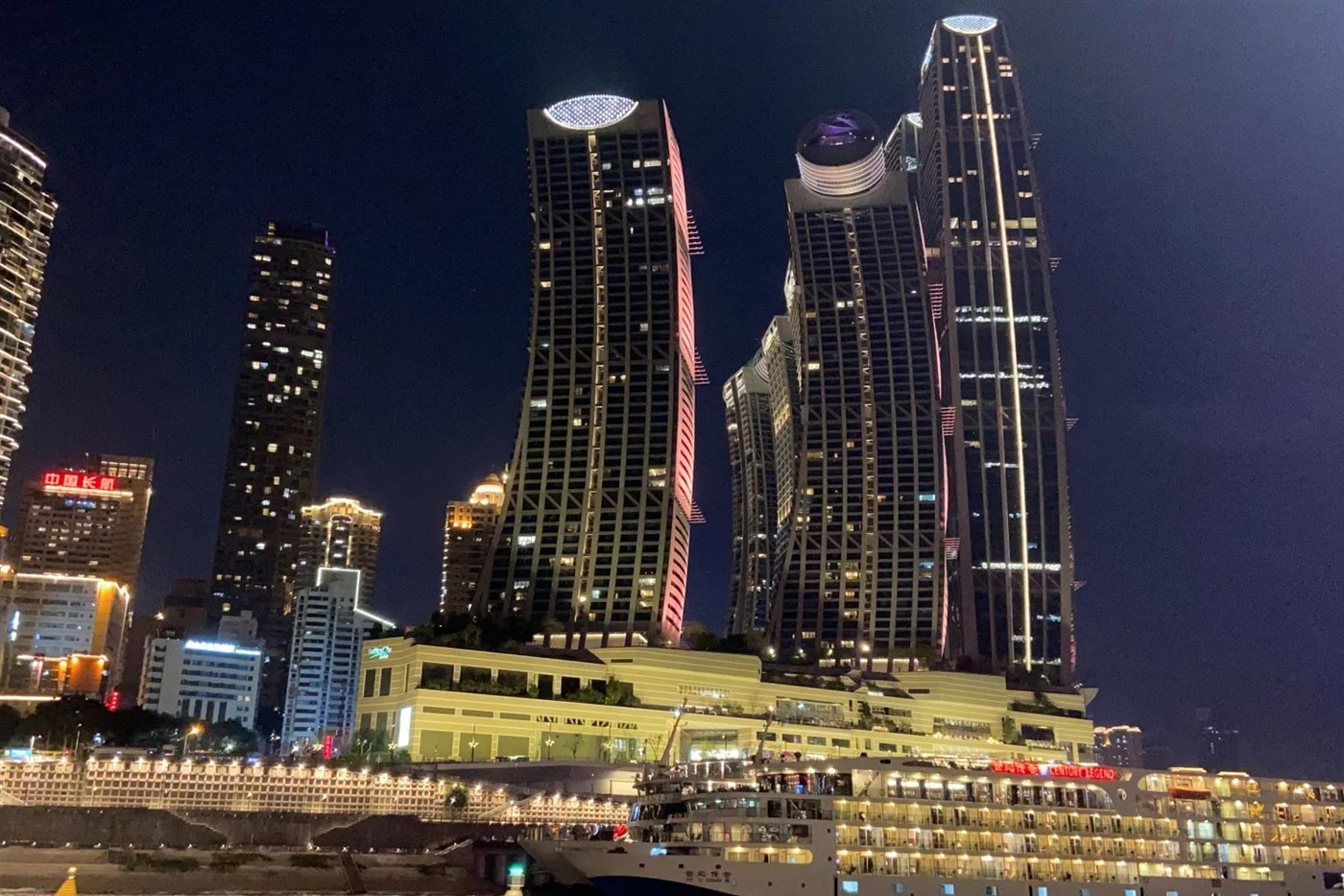 The Raffles City towers at Chaotianmen lit at night, seen from the water, with a large multi-deck river cruise ship named Century Legend berthed at the terminal below them.