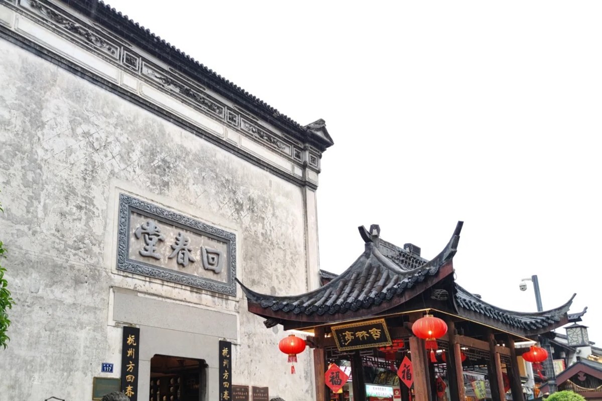The tall whitewashed gable of Fang Huichuntang pharmacy on Hefang Street in Hangzhou, carrying a carved stone plaque reading 回春堂, with a lantern-hung timber pavilion beside it in the rain.
