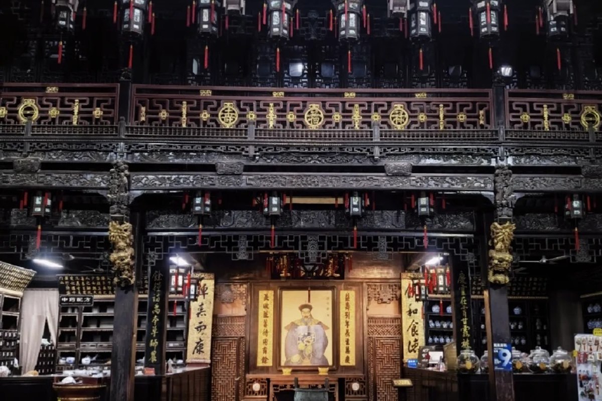 The double-height timber dispensing hall inside Huqing Yu Tang in Hangzhou — carved black-wood galleries, rows of hanging lanterns, walls of medicine drawers and a portrait of the founder at the far end.