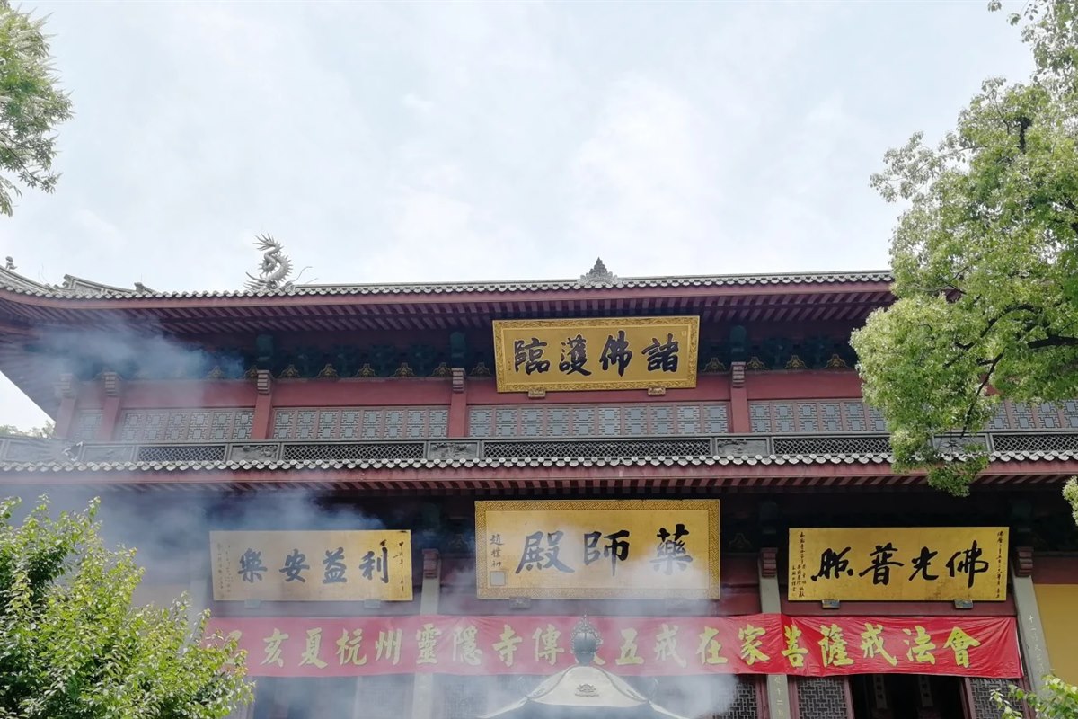 The Medicine Buddha Hall at Lingyin Temple in Hangzhou, its gold plaques half-veiled by drifting incense smoke, with a red banner across the front naming a temple ceremony.