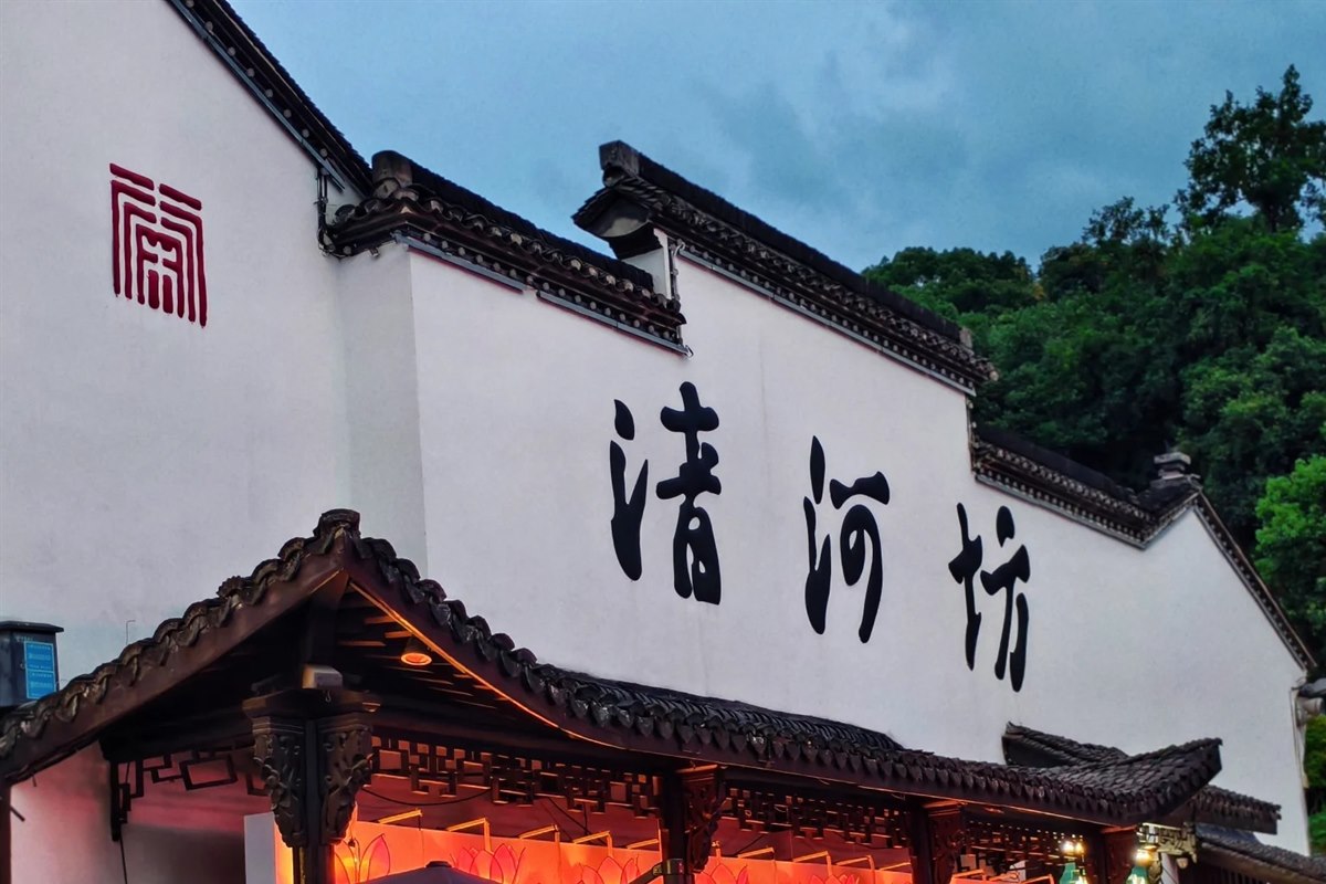 The white gable wall at the entrance to Qinghefang in Hangzhou, painted with the three large characters 清河坊, with lantern-lit shopfronts below and the wooded slope of Wushan hill behind.