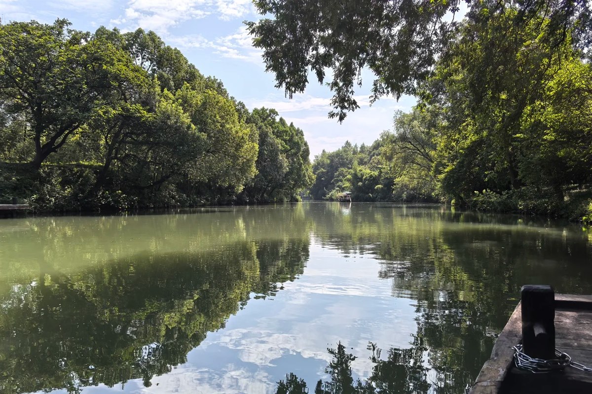 A tree-lined waterway at Xixi Wetland in Hangzhou seen from the bow of a boat, with still green water reflecting the canopy and another boat far ahead.