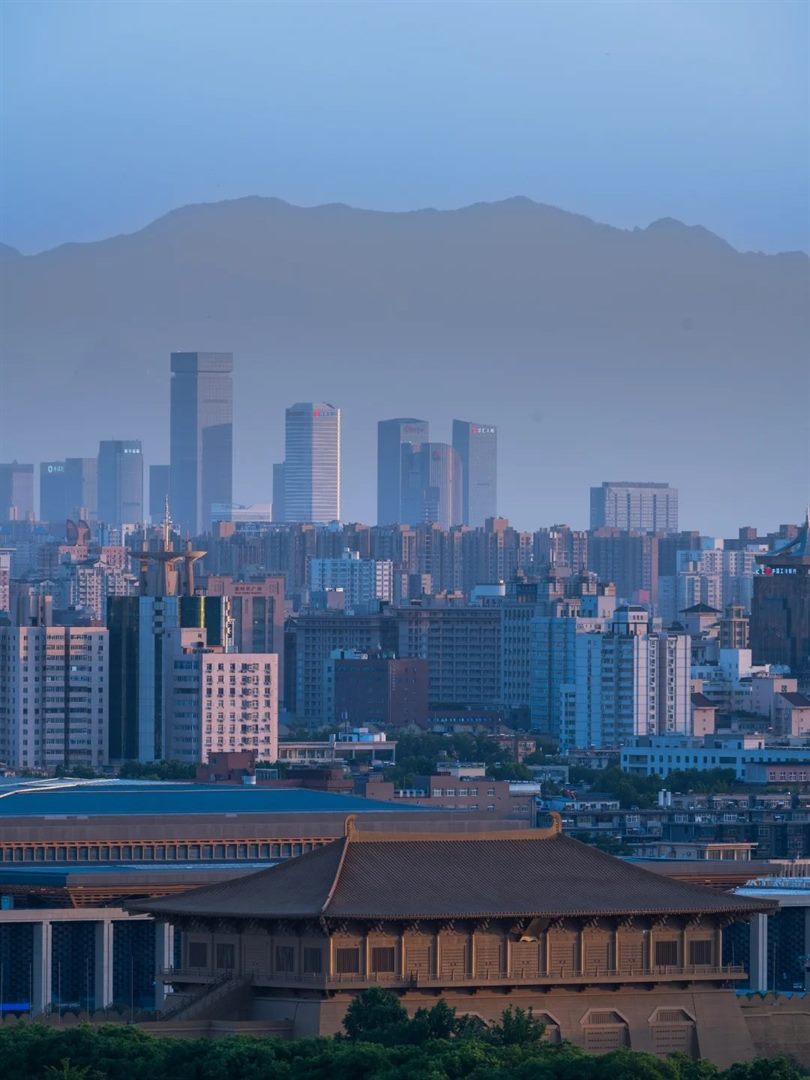 A dawn telephoto view stacking the hazy Qinling ridge, the towers of the Xi'an central business district, and the lit roof of Danfeng Gate sitting low among modern buildings.