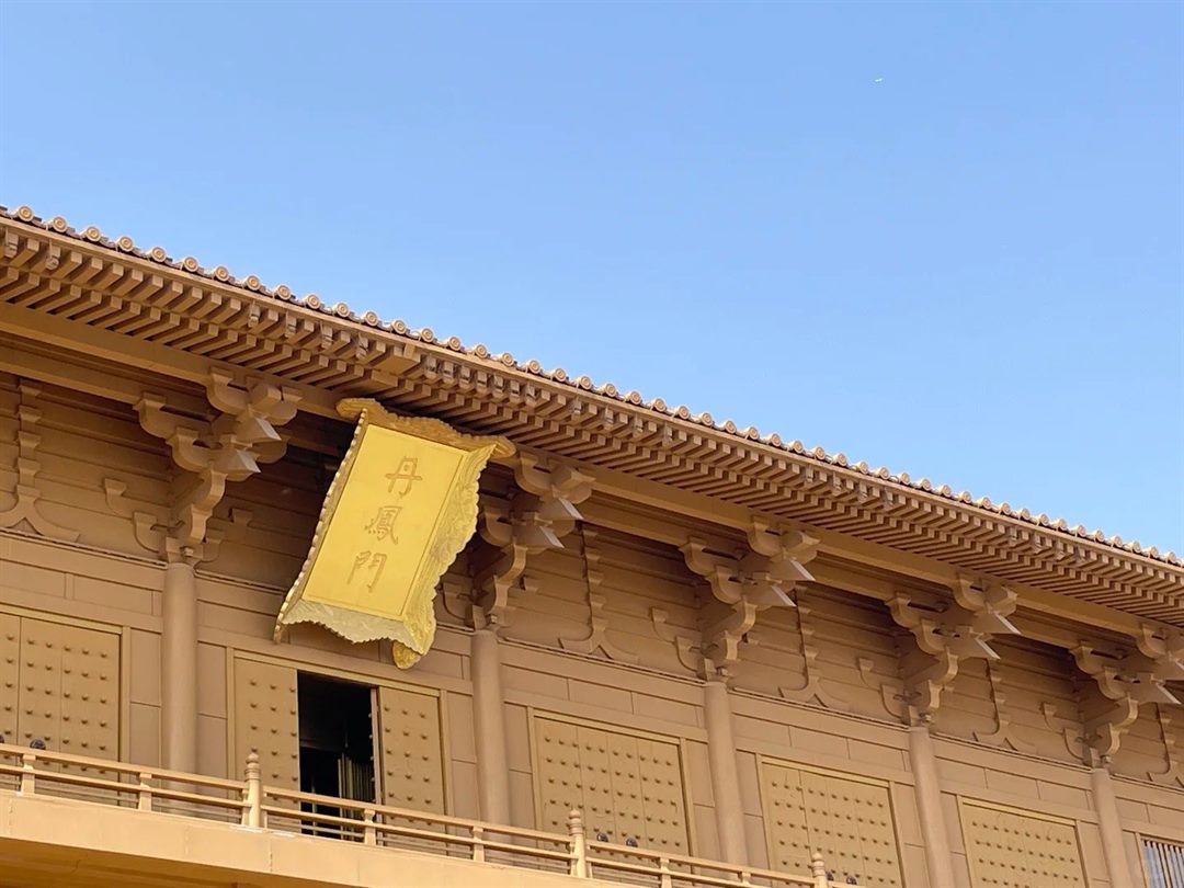 A raking close-up of unpainted ochre timber bracket sets and tile ends against a clear blue sky, with the gold 丹鳳門 plaque hanging at left.