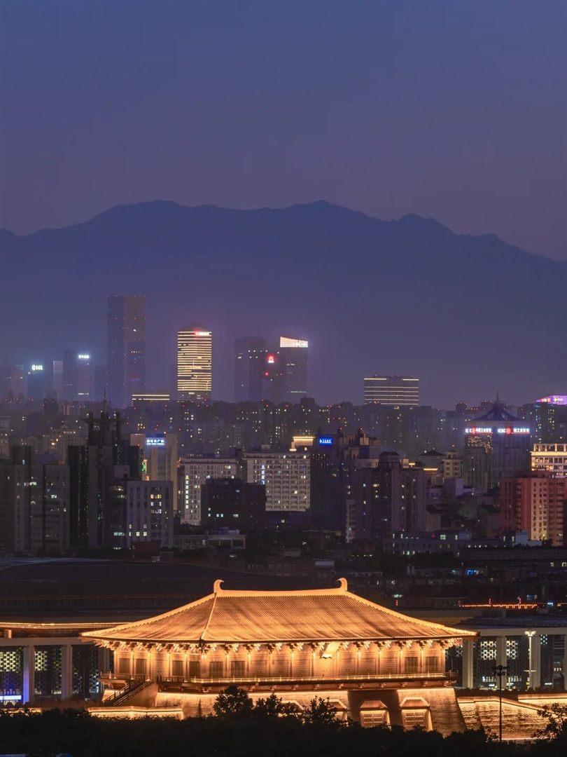 The same viewpoint at blue hour, with a purple ridge, the lit city towers, and Danfeng Gate floodlit amber among modern buildings.