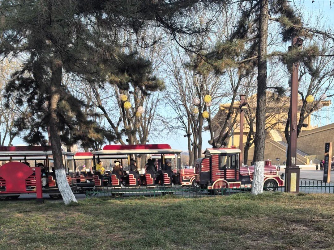 A red-and-cream land train with four open carriages of seated passengers, passing behind bare winter trees with the ochre Danfeng Gate rampart beyond.