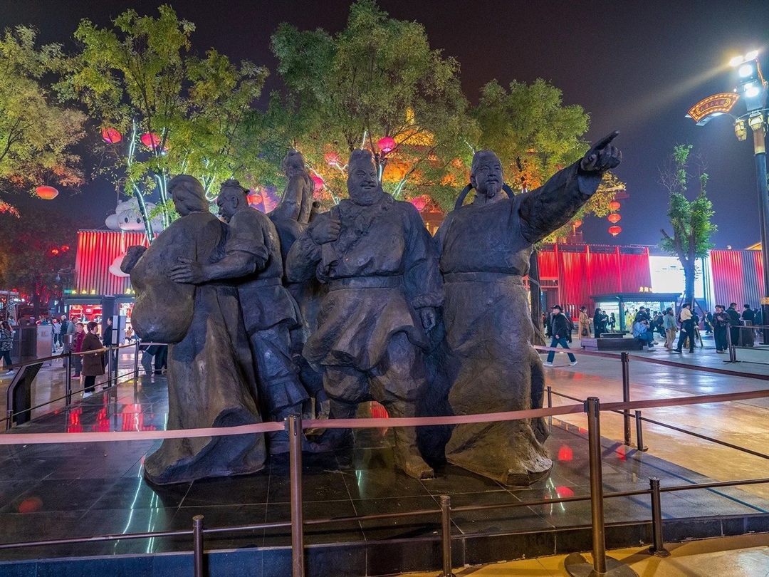 A bronze Silk Road merchant group on a railed plinth, one figure pointing, beneath trees strung with red lanterns, with lit shopfronts running back on both sides.