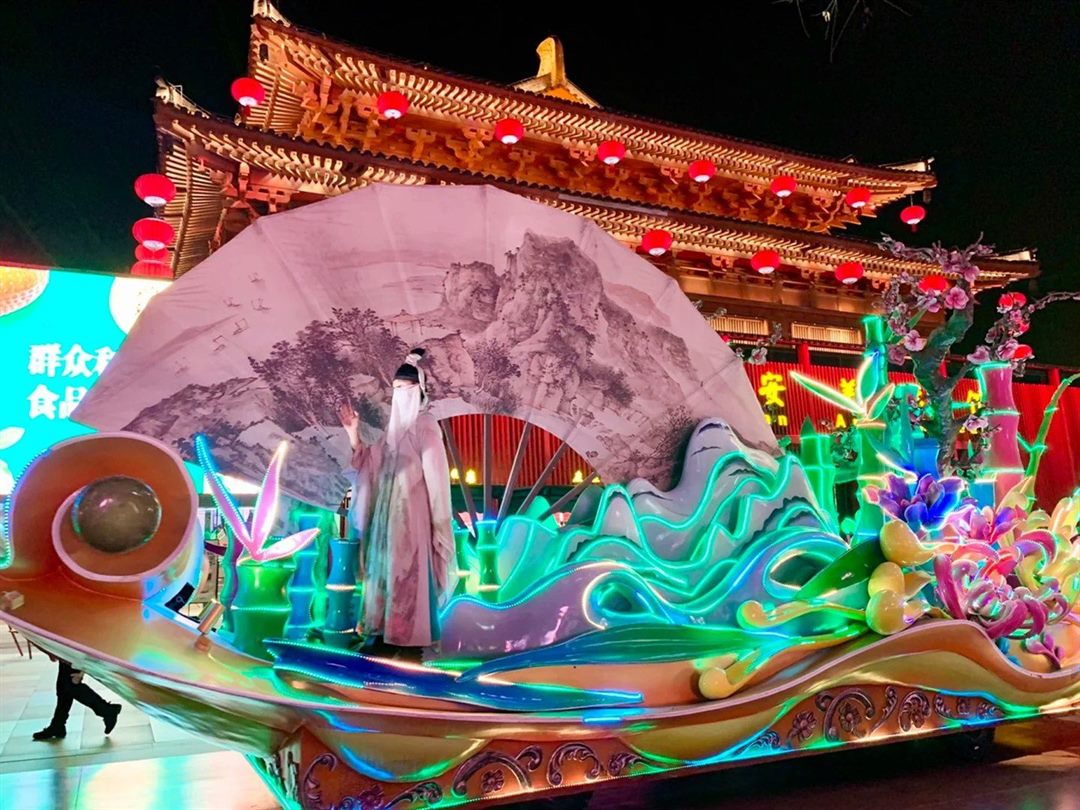 A giant painted-fan float with one veiled performer standing on the deck, hand raised, passing a Tang-style pavilion hung with red lanterns.