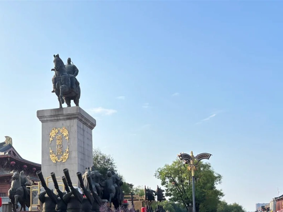 The same monument in flat daylight against a blue sky, with the gold plaque legible on the plinth and horn-bearer figures across the foreground.