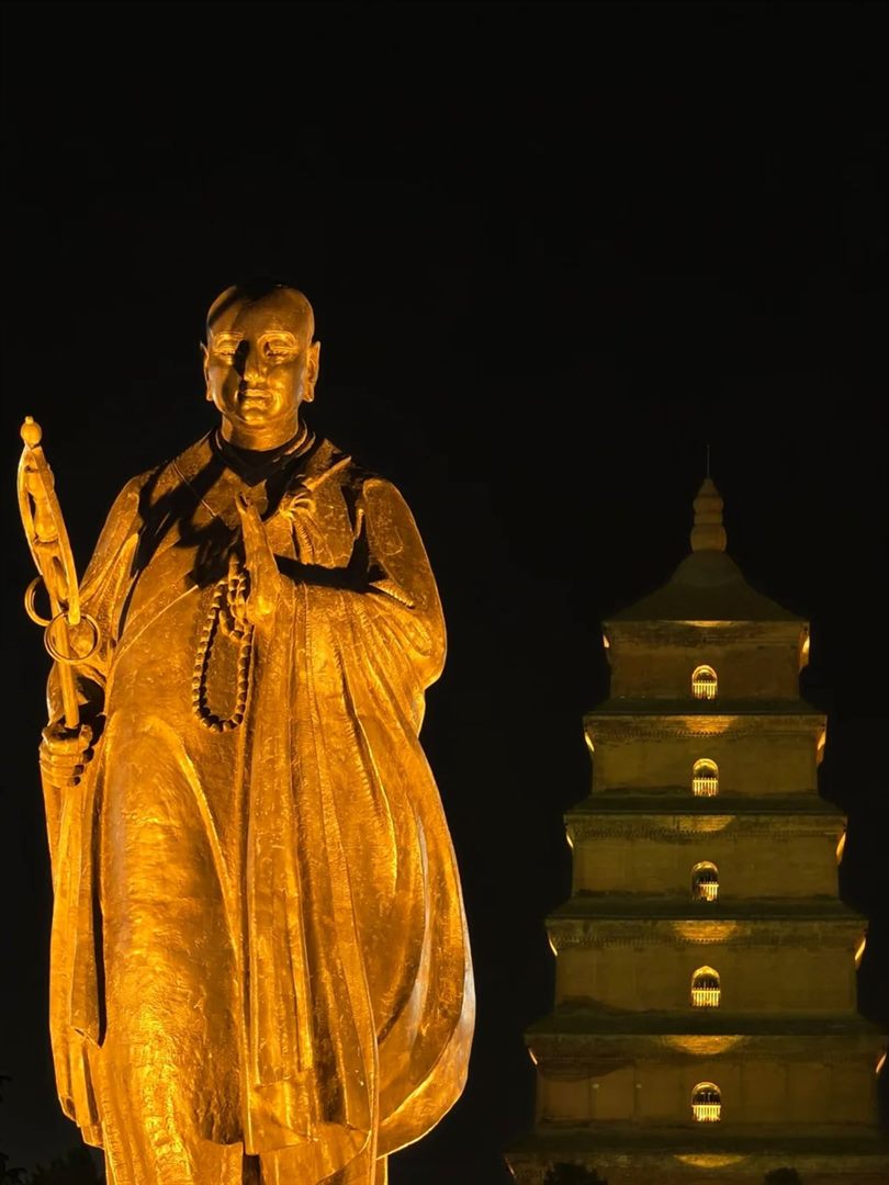 A gilded statue of the monk Xuanzang with staff and prayer beads, lit warm against a black sky, with the floodlit Big Wild Goose Pagoda rising behind to the right.