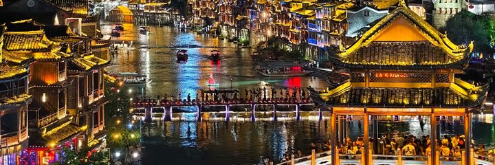 The lit Fenghuang riverfront at night seen from above, with boats on the Tuojiang and crowds crossing the stepping stones.