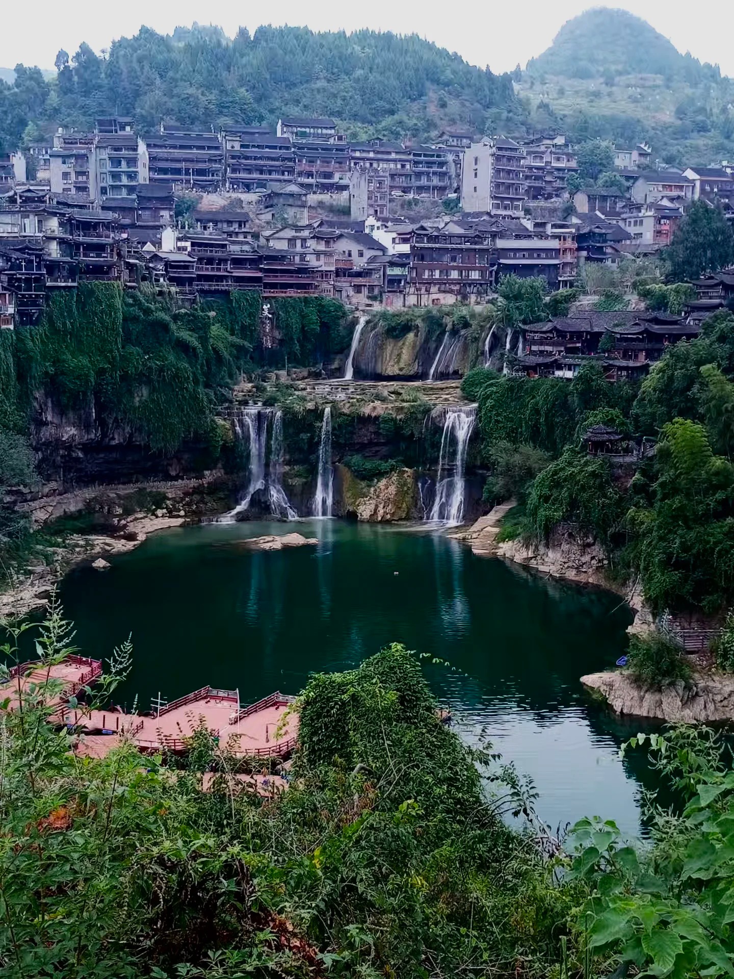 The Furong Ancient Town waterfall by day — multiple tiers dropping into an emerald plunge pool below the cliffside stilt houses.