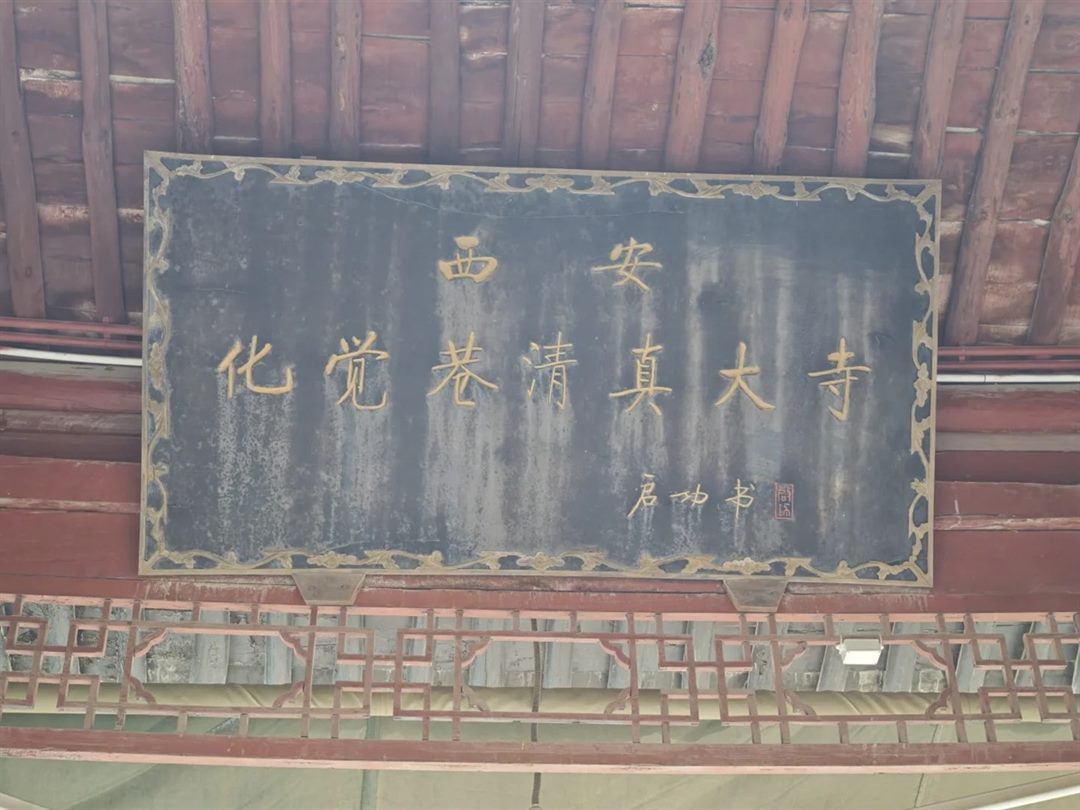 A dark name board under exposed red rafters, gold characters reading 西安 化觉巷清真大寺, signed 启功书 with a red seal.
