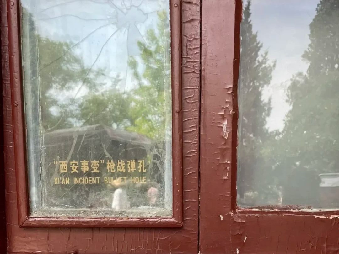 A window of the five rooms at Huaqing Palace with flaking red-brown paint, a bullet hole and radiating crack in the glass, and a gold bilingual label reading Xi'an Incident bullet hole.