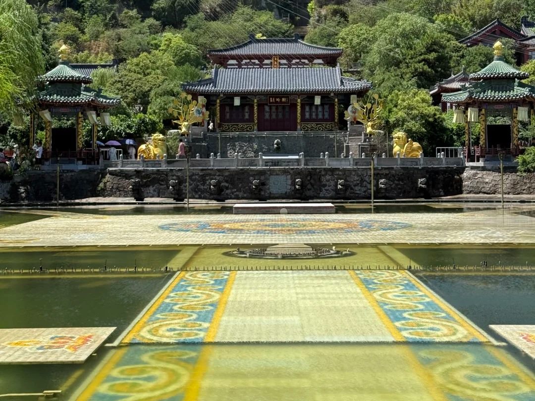 A broad tiled water terrace with decorative panels at Huaqing Palace leading to a hall flanked by gilded statues, with the wooded slope of Mount Li rising behind.