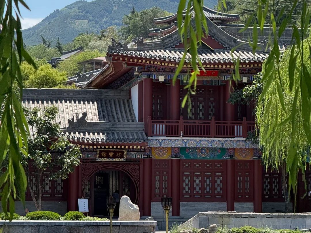 A red and white Tang-style hall with green tiled roofs at Huaqing Palace, framed by willow branches, with the wooded peak of Mount Li behind it against a blue sky.