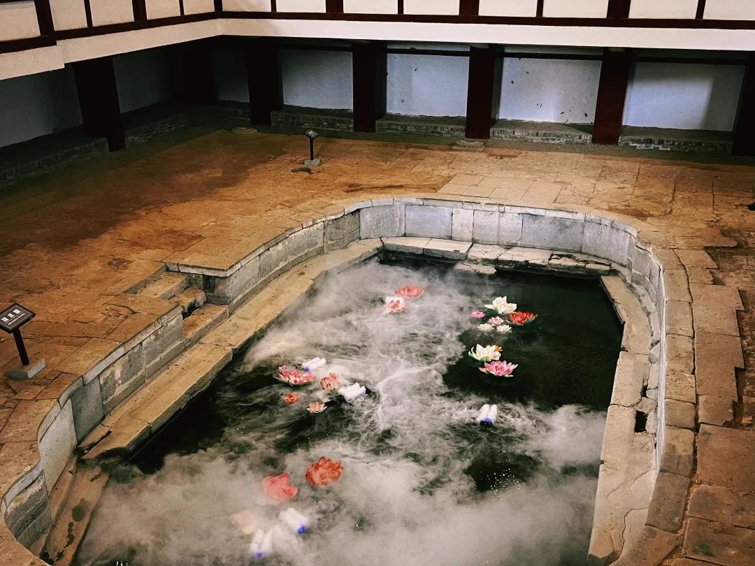 An excavated stone bathing pool sunk into a brick floor inside a covered hall at Huaqing Palace, with mist and artificial lotus flowers on the water and a display gallery above.