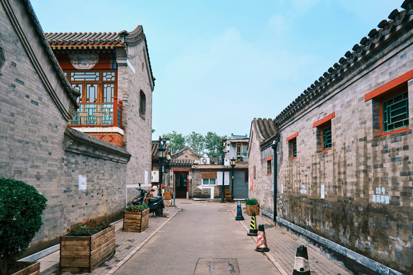 A quiet grey-brick hutong lane in Beijing lined with traditional courtyard-house walls, lattice windows and red trim.