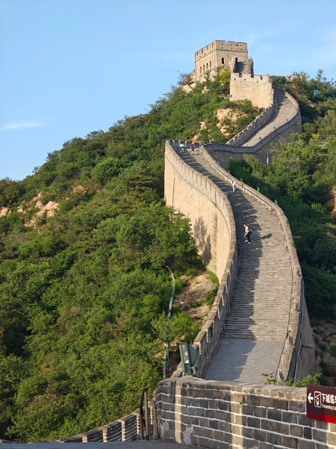 The Great Wall of China snaking across forested mountain ridges, a watchtower at the summit, visitors climbing the steps.