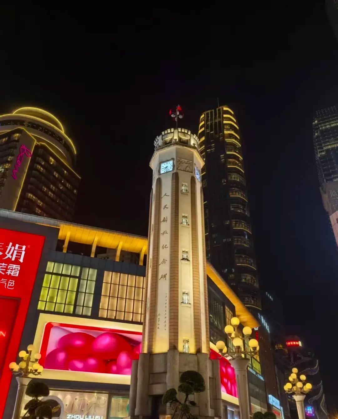 The People's Liberation Monument lit at night, its clock face and the vertical inscription visible, framed by the neon towers of the Jiefangbei CBD.