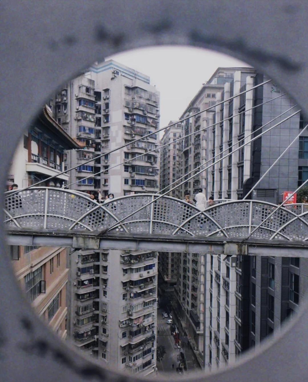 One of the Kuixinglou sky bridges seen through a circular opening: a white perforated-steel walkway with a few people on it, apartment towers behind and a street canyon far below.