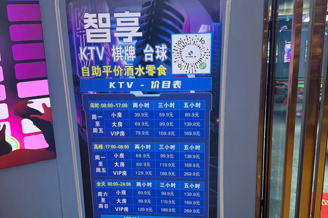 A KTV price signboard in a shopping centre, listing hourly rates by room size and time slot in Chinese yuan, next to a QR code for booking.