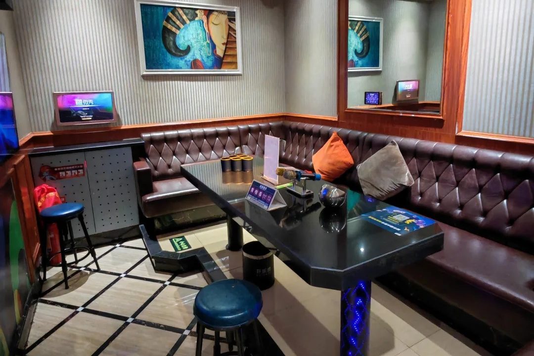 An ordinary 量贩式 KTV room: a leather sofa wrapping around a low table with two microphones, a small song-request screen mounted on the wall, and warm overhead lighting — no dim lounge lighting or rows of liquor bottles.