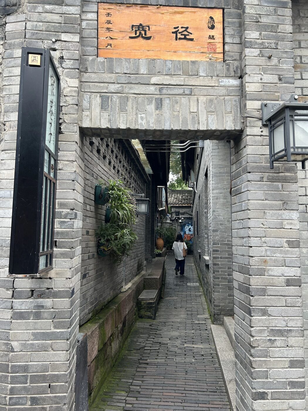 A narrow grey-brick lane at Kuanzhai Alley framed by a 宽径 plaque, with a visitor walking under planted courtyard walls, Chengdu.