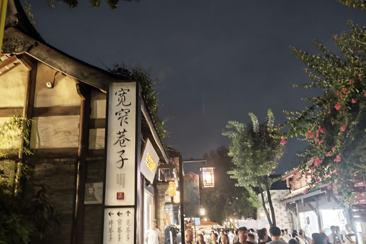 The Kuanzhai Alleys name sign and its three-way direction panel at night, with a lantern-lit lane and dense crowds behind.