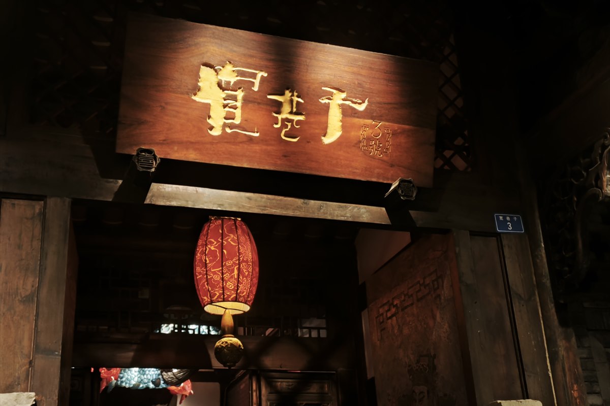 A carved wooden plaque reading 宽巷子 above a courtyard doorway on Wide Alley at night, a red lantern hanging beneath it.