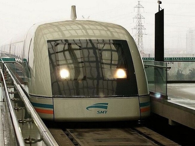 Shanghai Maglev train arriving at the platform — front-on view with headlights on, distinctive SMT-branded aerodynamic nose, white body with orange and turquoise stripes
