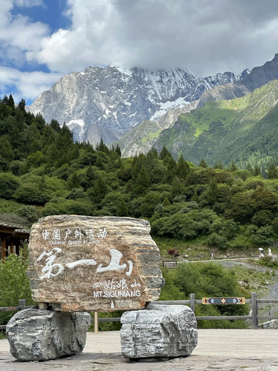 The stone marker at the Changping Valley trailhead, carved with 中国户外运动 第一山 and 四姑娘山 MT SIGUNIANG, with a snow-streaked rock face rising behind forested slopes.