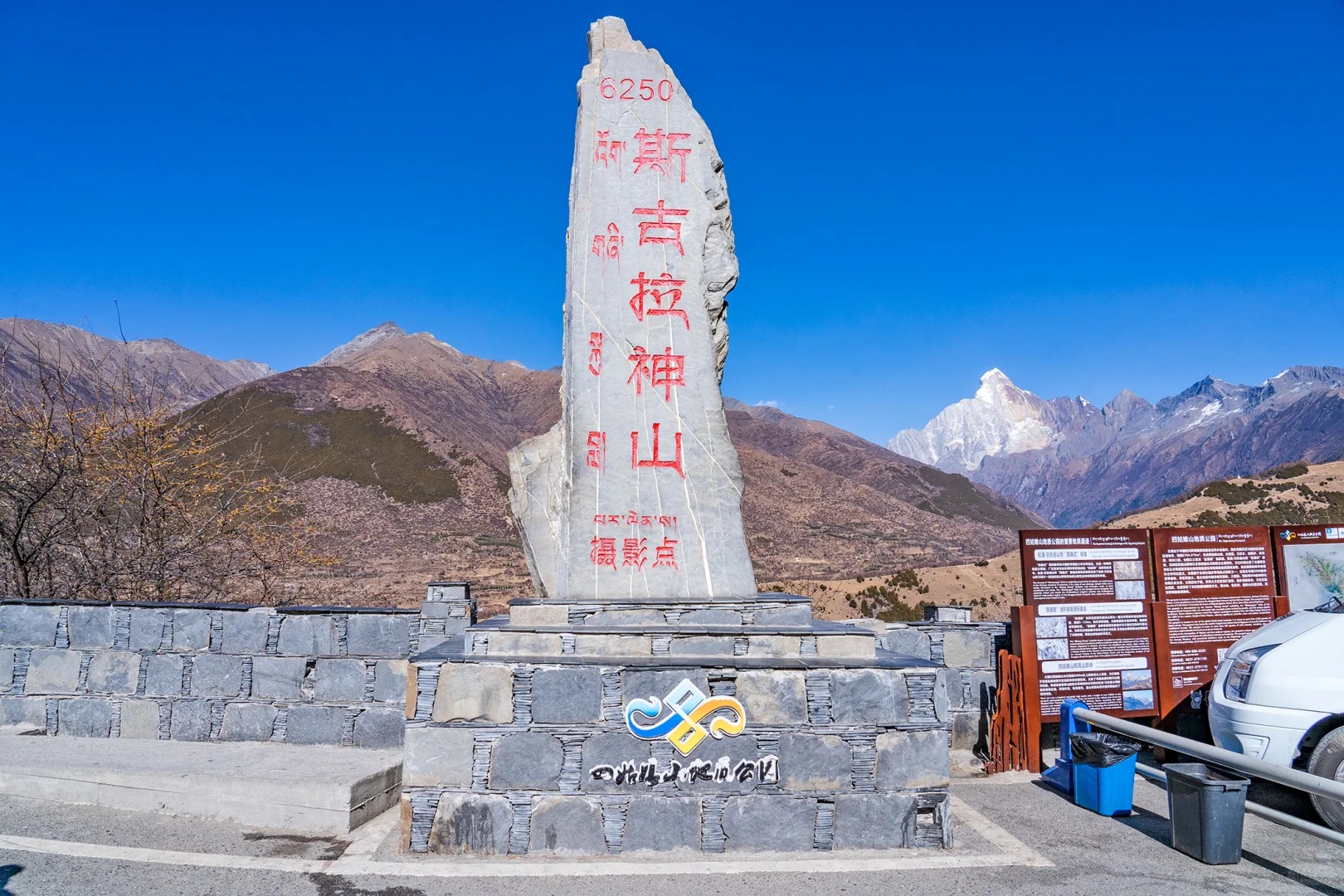 The Maobiliang roadside viewpoint: a tall inscribed monument on a plinth carrying the Mount Siguniang Geopark logo, a bank of trilingual interpretive boards and a park map, a steel guardrail, a parked car and bins, with the four snow summits visible over the ridge beyond.