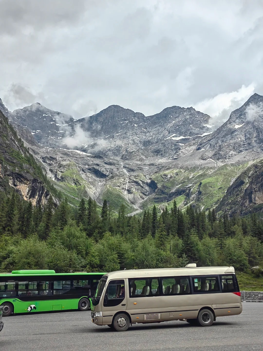 A beige coaster minibus and a green panda-liveried shuttle bus parked on a paved lot inside the scenic area, with glaciated peaks and a hanging glacier filling the background.