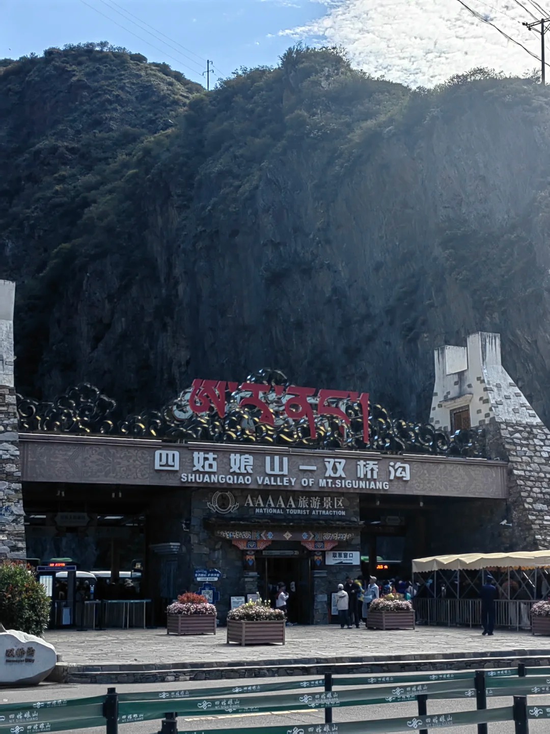 The Shuangqiao Valley entrance under a cliff: a sign reading 四姑娘山—双桥沟 SHUANGQIAO VALLEY OF MT.SIGUNIANG with a 5A plaque, a ticket-collection window, a queue canopy and turnstiles, with visitors crossing the plaza.