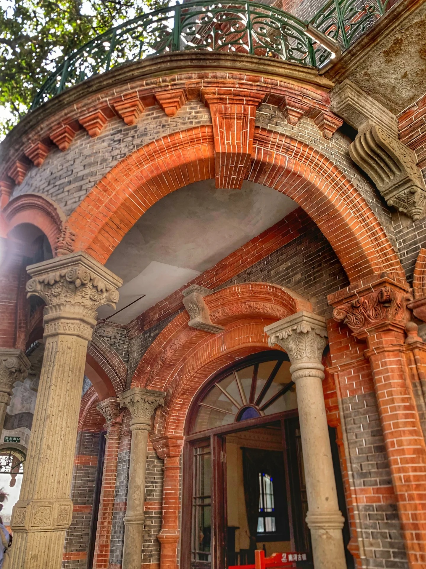 A two-storey arcade in a Nanxun merchant mansion: bright red brick arches springing from carved pale-stone Corinthian columns, with grey brick infill behind and a fanlight window over the doorway.