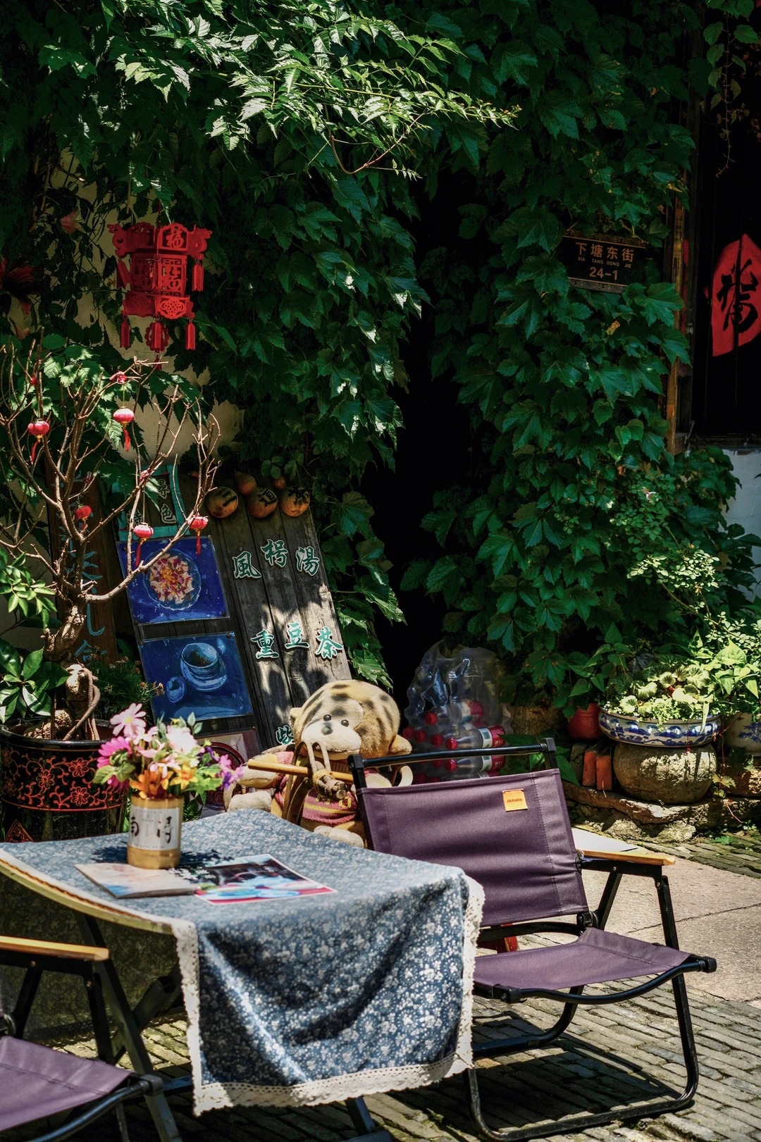 A teahouse's outdoor tables on a Nanxun lane: folding chairs and a blue floral cloth under a wall of creeper, with a painted board advertising fengxiao soup and smoked-bean tea, and a bilingual street plate reading Xia Tang Dong Jie 24-1 above.