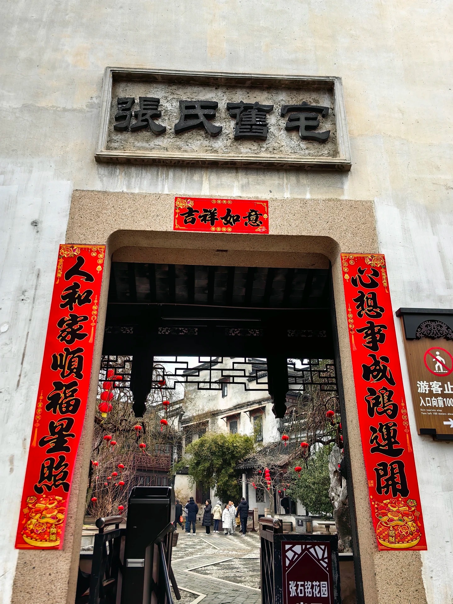 The entrance to the Zhang family's former residence in Nanxun: a plain rendered wall with a carved stone-framed plaque reading Zhang Shi Jiu Zhai, red New Year couplets down each side of the doorway, and a lantern-hung courtyard beyond.