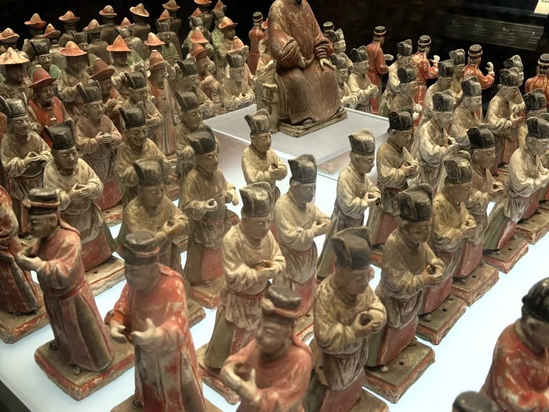 A dense array of small painted pottery figures on a lit plinth in a glass case at the Shaanxi History Museum, with a larger seated figure behind them.