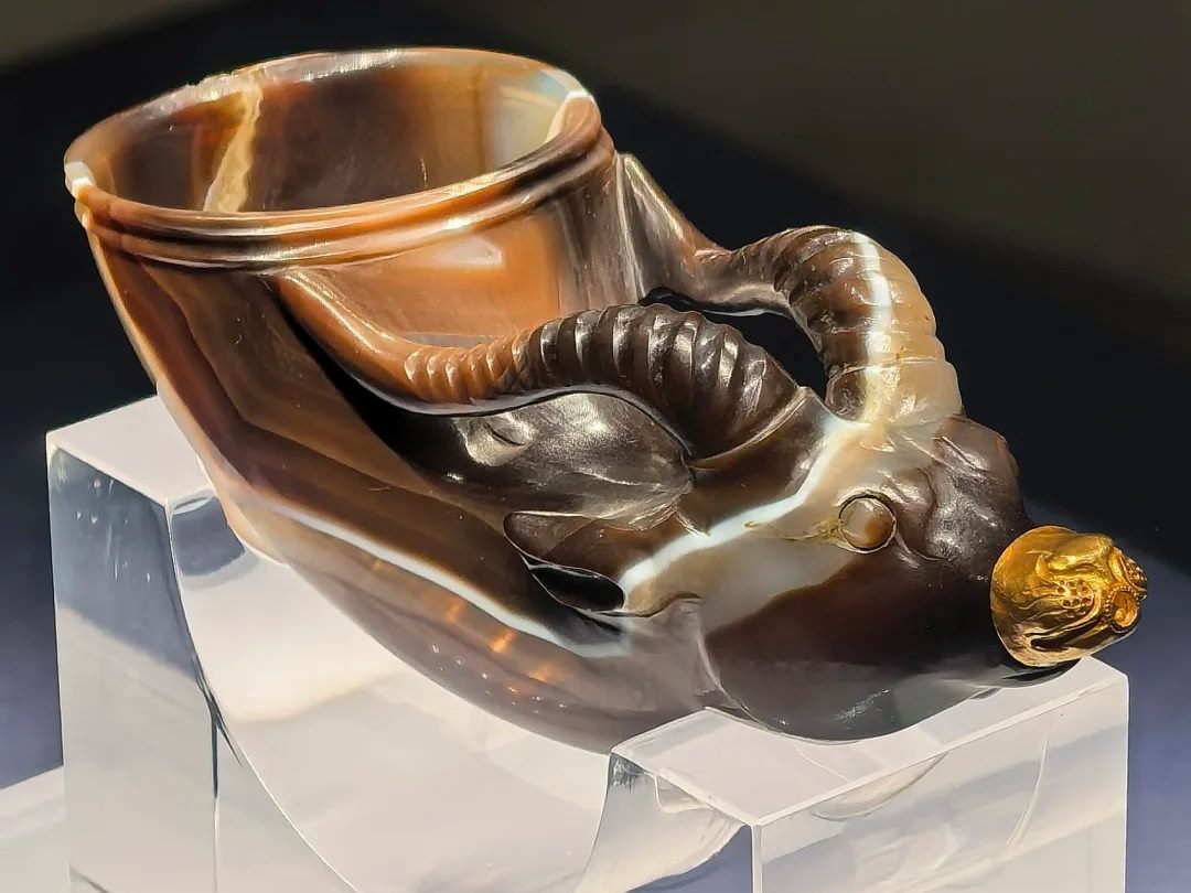 A banded agate drinking vessel carved as an animal's head, with a gold muzzle cap, on a clear acrylic stand in a dark display case.