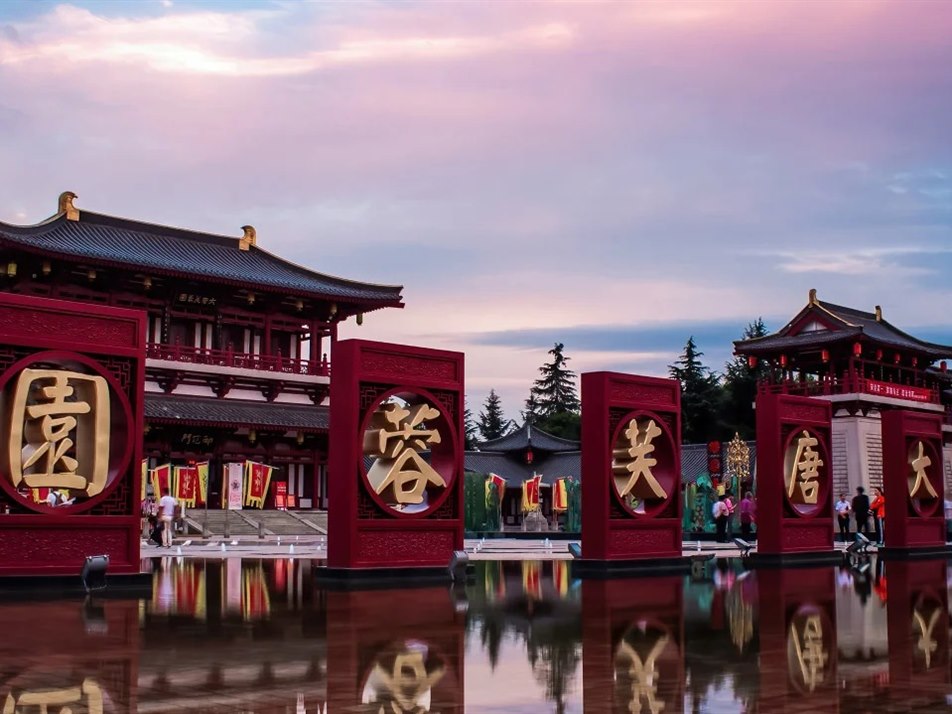Five red character-frames reading 大唐芙蓉園 standing in a shallow mirror pool and reflected in it, with the gate building behind under a mauve dusk sky.