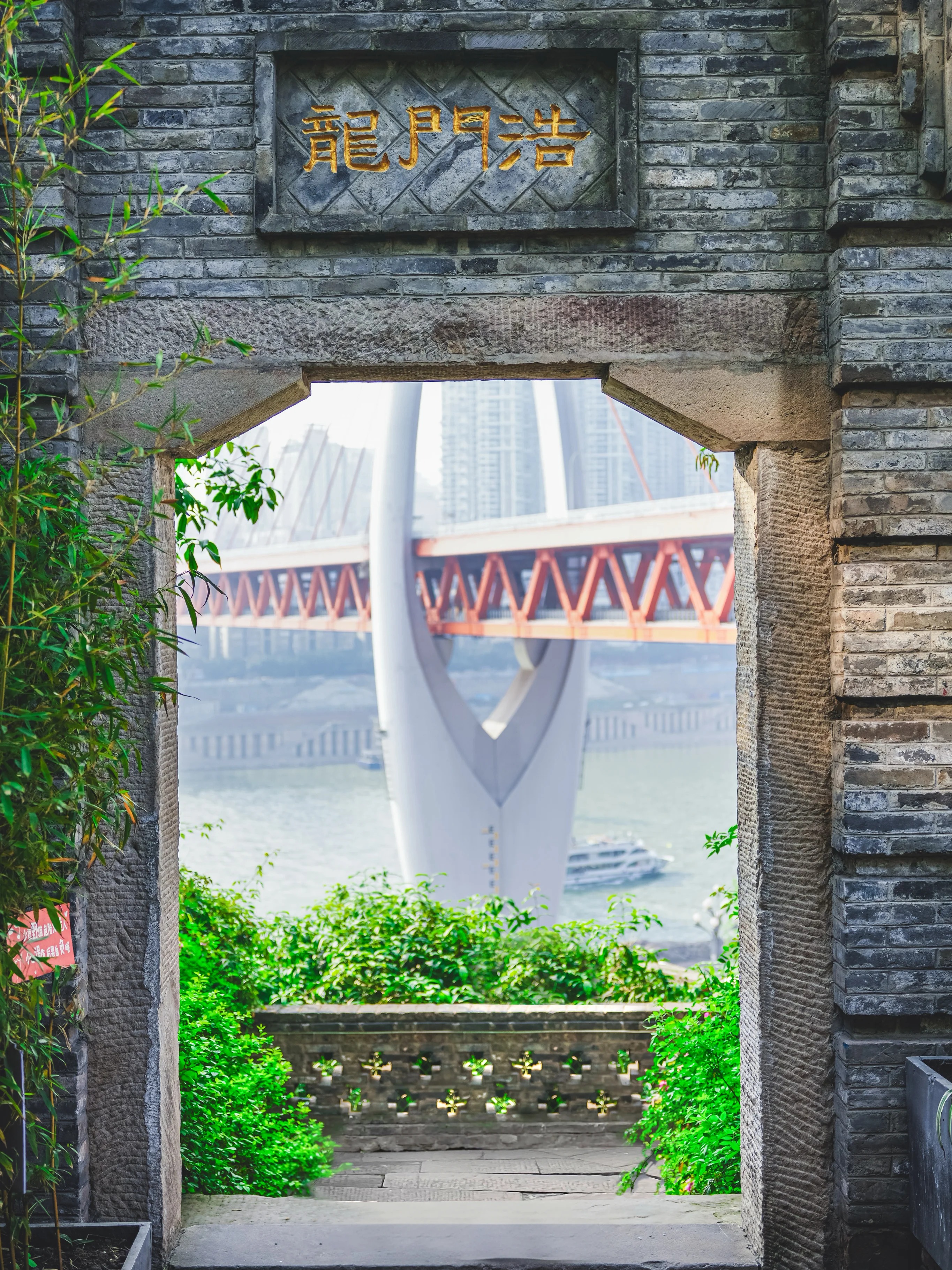 A grey brick gateway with 龍門浩 carved in gold on the lintel, framing a view through to the white pylon and orange truss deck of Dongshuimen Bridge, with a cruise boat on the river below.